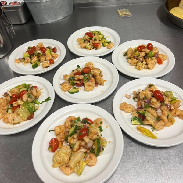Seven plates of Cajun shrimp salad with tomatoes, cucumbers, and onions on a metal counter.